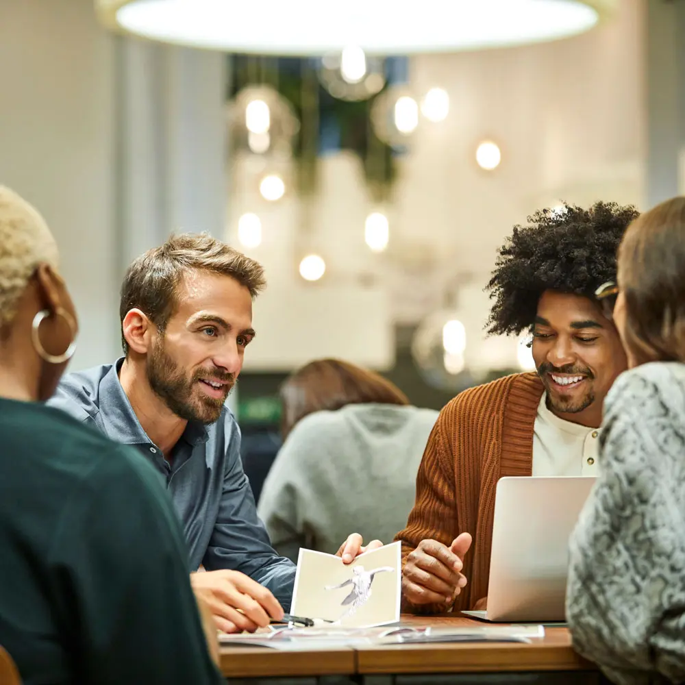 Grupo de personas sentadas alrededor de una mesa, conversando y trabajando en una presentación. Uno de los hombres sonríe mientras sostiene un objeto en la mano. Hay documentos y una computadora portátil sobre la mesa. El ambiente es moderno y acogedor.
