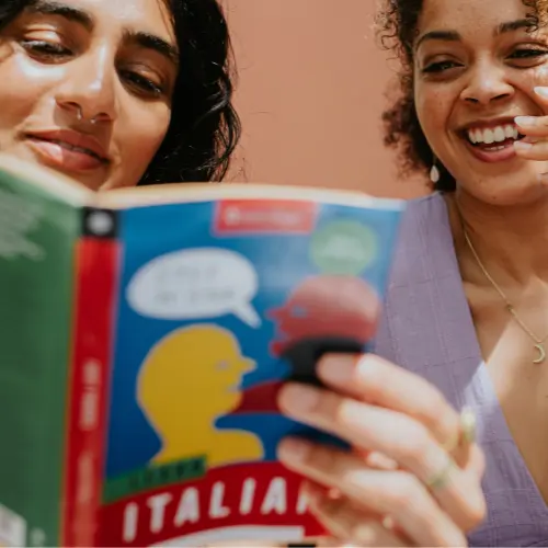 Dos mujeres jóvenes se ríen mientras leen un libro titulado "Italiano". Una de ellas sostiene el libro, mostrando una ilustración colorida en la portada. Ambas parecen disfrutar de la lectura y comparten un momento divertido juntas. El fondo es de color liso.
