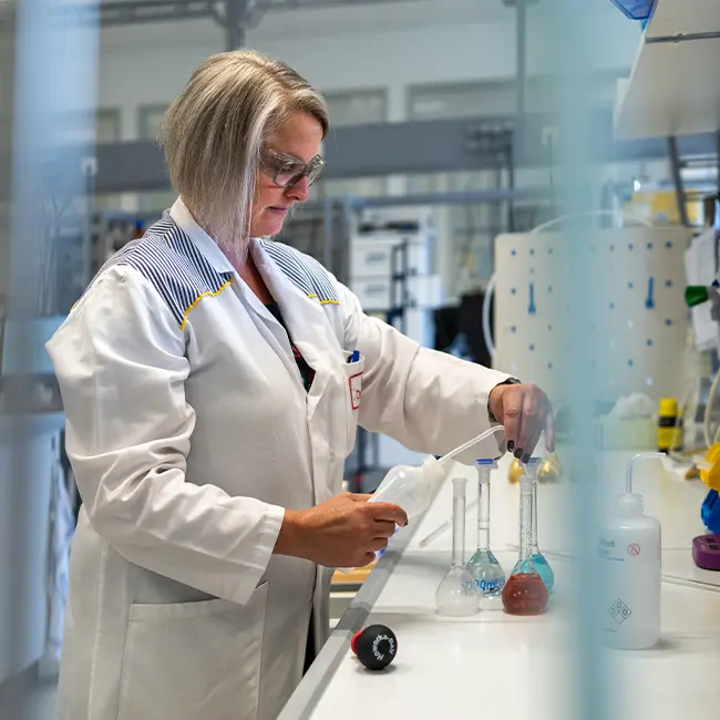 Una mujer con gafas y un lab coat blanco trabaja en un laboratorio, manipulando frascos de vidrio y equipos de medición. El entorno es de un laboratorio científico, con herramientas y sustancias químicas visibles sobre la mesa.