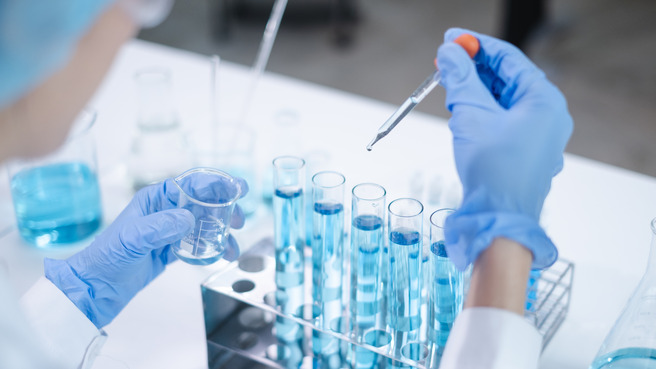 Manos con guantes azules sosteniendo un tubo de ensayo y una pipeta, mientras se preparan para añadir un líquido azul a varios tubos de ensayo alineados. En el fondo, hay frascos y material de laboratorio.
