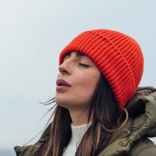 Mujer con gorro naranja y abrigo, con los ojos cerrados y una expresión de tranquilidad en su rostro, parece disfrutar de un momento de paz al aire libre. El fondo es difuso, sugiriendo un ambiente abierto y natural.