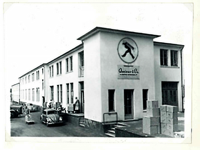 Edificio de una empresa con un gran logotipo en la fachada. Se observan personas y vehículos estacionados frente a la entrada. Cajas apiladas en el lado derecho. La imagen tiene un estilo envejecido y es en blanco y negro.