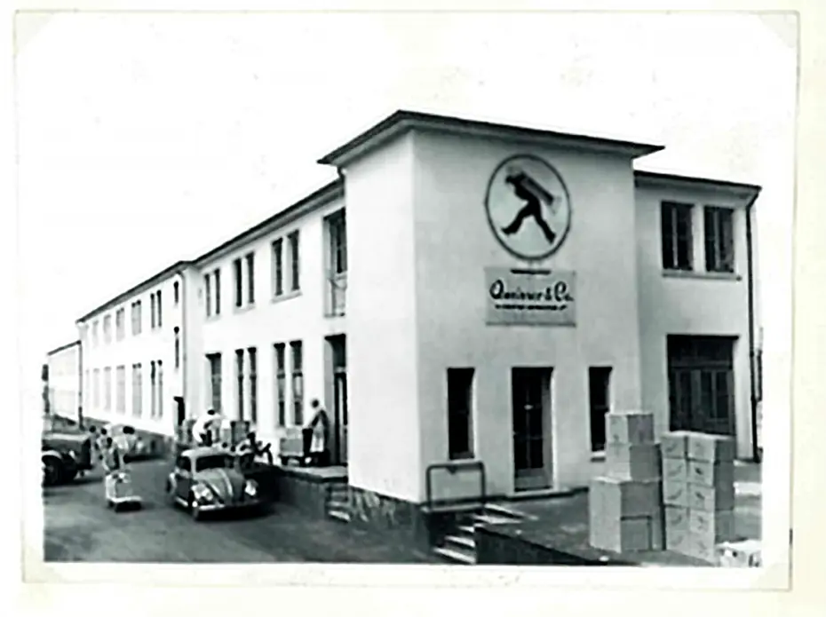 Edificio de una empresa con un gran logotipo en la fachada. Se observan personas y vehículos estacionados frente a la entrada. Cajas apiladas en el lado derecho. La imagen tiene un estilo envejecido y es en blanco y negro.