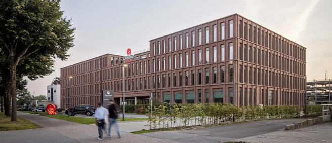 Edificio moderno de ladrillo con grandes ventanas, ocupado por un negocio. Se observan dos personas caminando por la acera frente al edificio, y vehículos estacionados. Al lado, hay una escultura roja y árboles que rodean el entorno.