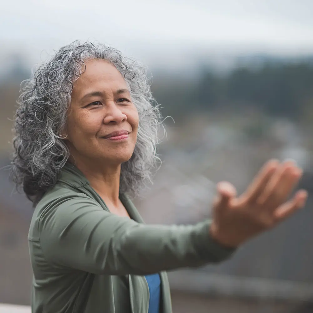 Mujer mayor con cabello rizado y canoso, sonriendo y extendiendo una mano hacia adelante. Viste una chaqueta gris y un top azul. De fondo, un paisaje urbano con un cielo nublado.