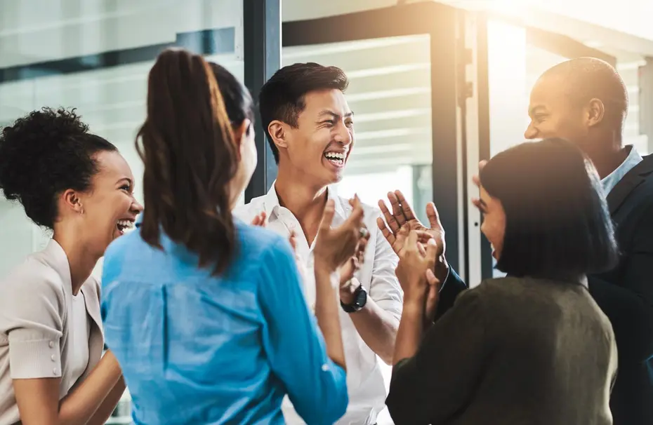 Grupo diverso de seis personas riendo y aplaudiendo en un ambiente alegre y profesional. Están interactuando de manera entusiasta, mostrando sonrisas y camaradería. La luz natural entra por una ventana, creando un ambiente cálido y positivo.