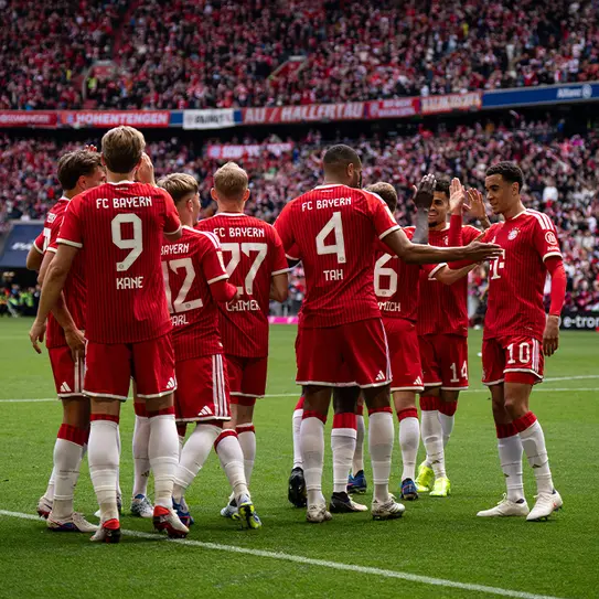 Jugadores del FC Bayern celebrando en el campo, vistiendo camisetas rojas con blanco. Se agrupan para una charla motivacional antes del partido, con el público de fondo animando.
