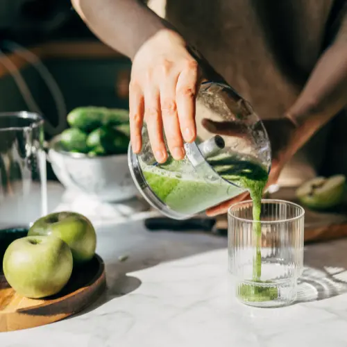 Una mano vierte un batido verde en un vaso transparente, mientras se ven manzanas verdes y pepinos en la mesa. La luz natural ilumina la escena, resaltando la frescura de los ingredientes. 