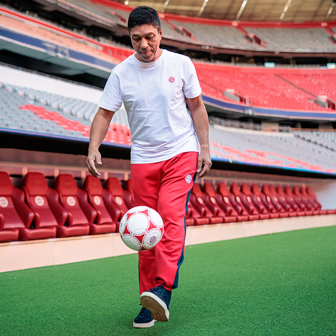 Un hombre sonriente viste una camiseta blanca y pantalones rojos, driblando un balón de fútbol en un estadio vacío. El césped está bien cuidado y se pueden ver las gradas con asientos rojos detrás de él.
