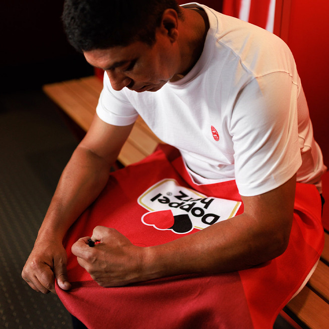 Hombre sentado en un vestuario, vistiendo una camiseta blanca. Está ajustando una toalla roja con un diseño que dice "Dopet Piz". El fondo muestra bancos de madera y paredes rojas.