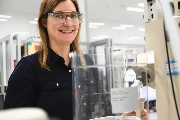 Mujer sonriente con gafas protectoras en un laboratorio, frente a un equipo de laboratorio. La imagen muestra un entorno limpio y moderno, con equipos de laboratorio al fondo.