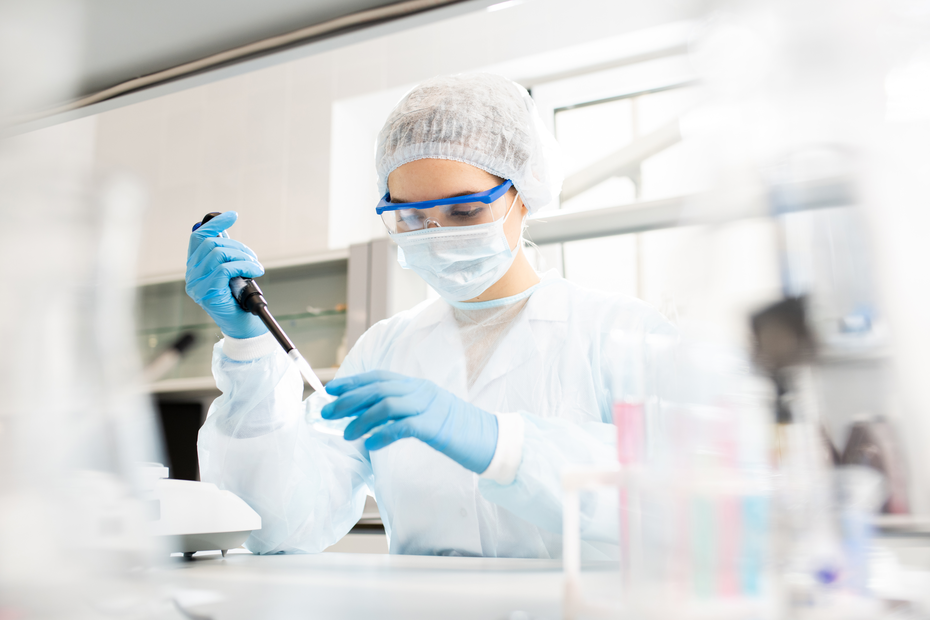 Una persona en un laboratorio, vestida con un equipo de protección que incluye bata, guantes y mascarilla, está utilizando una micropipeta. El entorno se ve limpio y ordenado, con varios utensilios de laboratorio visibles.