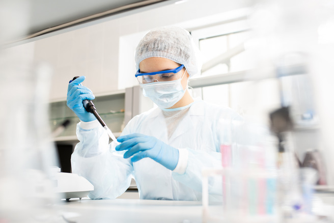Una persona en un laboratorio, vestida con un equipo de protección que incluye bata, guantes y mascarilla, está utilizando una micropipeta. El entorno se ve limpio y ordenado, con varios utensilios de laboratorio visibles.