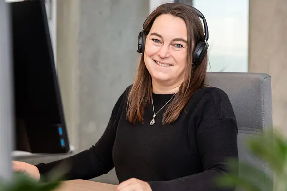 Mujer sentada frente a un escritorio, sonriendo mientras usa auriculares. Hay un monitor de computadora visible detrás de ella. La mujer tiene el cabello largo y castaño y viste un suéter negro. En el fondo, se pueden ver elementos de oficina y plantas.