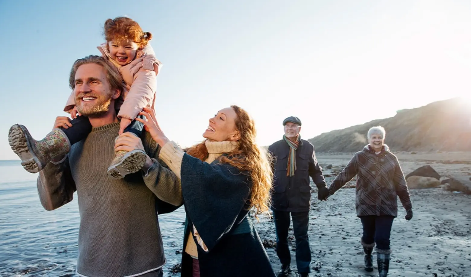 Una familia disfruta de un día en la playa. Un hombre lleva a una niña a hombros, mientras una mujer sonríe a su lado. En el fondo, dos adultos mayores caminan tomados de la mano. La escena transmite alegría y unión familiar.