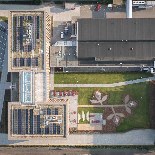 Vista aérea de un edificio moderno con techos solares, rodeado de áreas verdes y caminos. Se pueden ver espacios al aire libre con estructuras de diseño en el paisaje, así como estacionamiento en la parte inferior de la imagen.