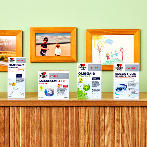 En la imagen se presentan varios productos en una estantería de madera: Omega-3 líquido, Magnesio 400 Citrato, Omega-3 vegetal y Augen Plus. Al fondo hay tres cuadros con dibujos infantiles y una fotografía con niños. Las paredes son de color verde claro.
