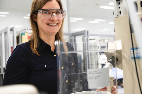 Mujer sonriente con gafas protectoras en un laboratorio, frente a un equipo de laboratorio. La imagen muestra un entorno limpio y moderno, con equipos de laboratorio al fondo.