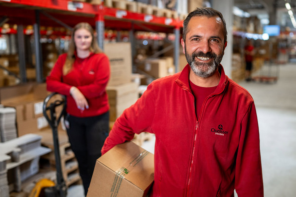 Dos empleados en un almacén están trabajando. Un hombre sonriente lleva una caja en la mano, mientras que una mujer de pie detrás de él observa. Ambos visten ropa roja y hay estanterías llenas de cajas en el fondo.