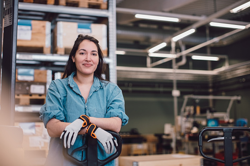Mujer con guantes de trabajo sonriente, apoyada en un transpaleta en un entorno industrial con estanterías llenas de cajas de madera y equipos en el fondo.
