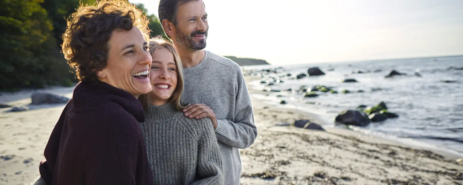 Tres personas sonrientes están de pie en la playa, con el mar y rocas visibles al fondo. La mujer a la izquierda lleva un abrigo oscuro, mientras que la joven en el centro usa un suéter claro. El hombre a la derecha lleva un suéter gris claro.